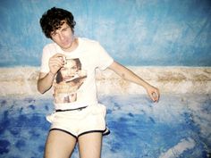 a young man in white shirt and shorts posing for the camera with blue wall behind him