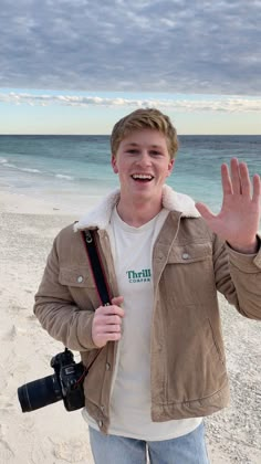 a young man standing on top of a sandy beach holding a camera and making a hand gesture