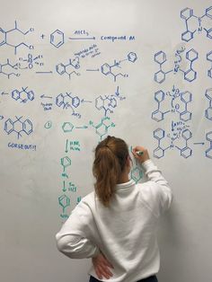 a woman writing on a whiteboard with blue marker and chemical diagrams drawn on it