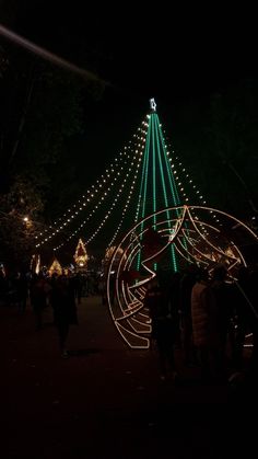 people are walking around in front of a christmas tree with lights on it at night