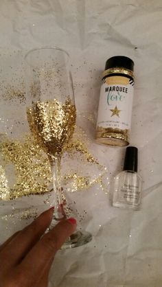 a hand holding a wine glass with gold glitter next to it and a bottle of marquee love