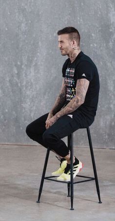 a man sitting on top of a black chair in front of a cement wall with his legs crossed