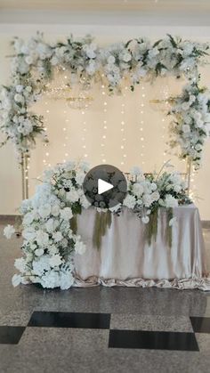 a table topped with white flowers and greenery next to a wall covered in lights