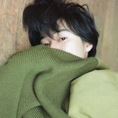 a man with black hair covering his face behind a green sweater and leaning against a wooden wall