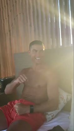 a shirtless man sitting on a bed with his hand in his pocket and looking at the camera