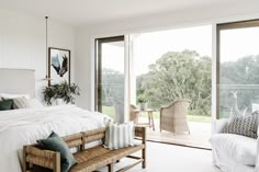 a bedroom with white walls and flooring has a large bed, wicker bench, and sliding glass doors leading to an outside deck area