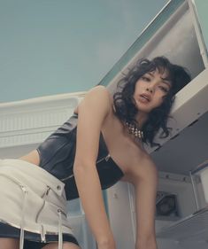 a woman in black top and white skirt leaning on a refrigerator door with her legs crossed