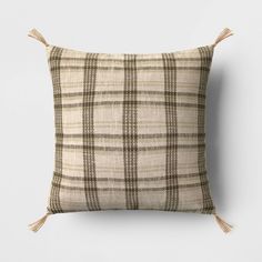 a beige and black plaid pillow with tassels on the sides, sitting against a white wall