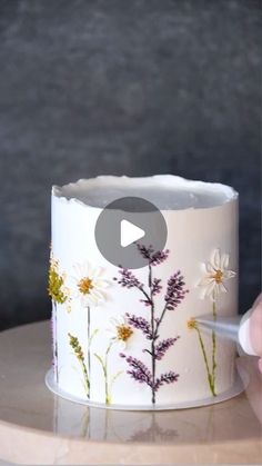 a white cake with flowers painted on it and a person holding a plastic object in front of the cake