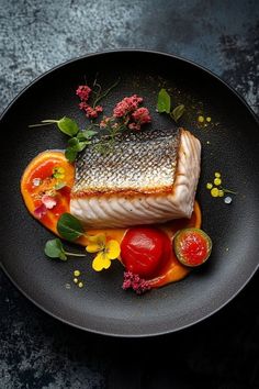 a black plate topped with fish and vegetables on top of a dark table next to utensils