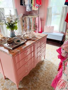 a pink dresser with lots of clothes on it in a room filled with windows and curtains