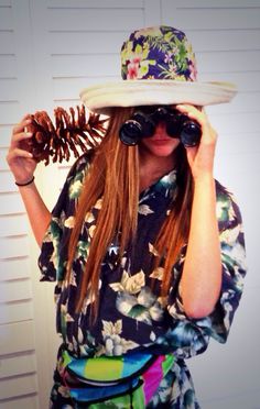 a woman with long hair wearing a hat and looking through binoculars while holding a pine cone