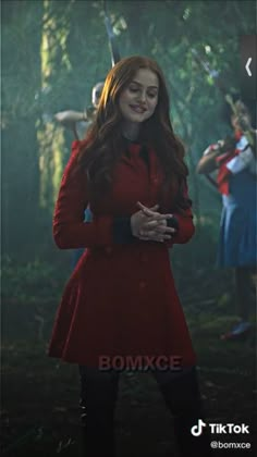 a woman in a red coat and black boots standing in the woods with her hands on her hips