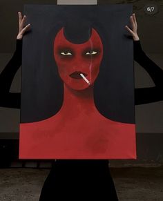 a woman holding up a painting with yellow eyes and red devil makeup on it's face