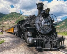 an old fashioned train is parked on the tracks in front of some mountains and clouds