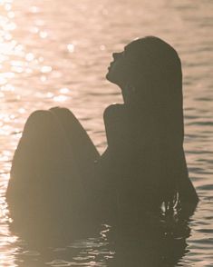 a woman sitting in the water with her back turned to the side, looking up at the sun