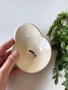 a hand is holding a small bowl next to some green leaves and sprigs