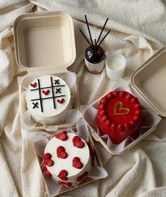 three cakes in plastic containers on a bed with white sheets and one has a red heart