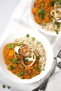 two bowls filled with rice and vegetables on top of a white cloth next to silverware