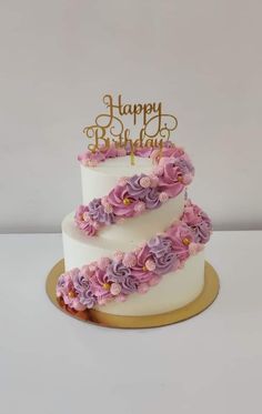 a three tiered white cake with purple flowers on the bottom and gold lettering happy birthday