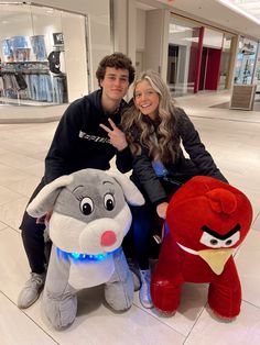 a man and woman pose with stuffed animals in an airport lobby, one is giving the peace sign