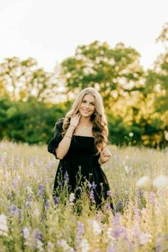 a woman standing in the middle of a field with purple flowers and trees behind her