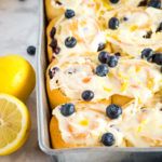 lemon blueberry rolls with icing and fresh blueberries in a baking pan next to sliced lemons