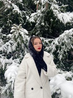 a woman standing in front of snow covered trees wearing a white coat and black scarf