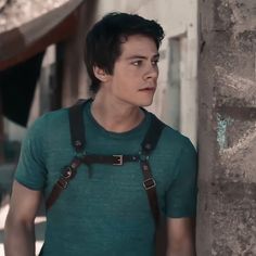 a young man with suspenders and a green t - shirt standing next to a stone wall