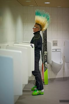 a man with green hair standing in a bathroom next to urinals wearing skis