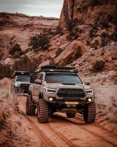 two trucks driving down a dirt road in the desert with rocks and grass behind them