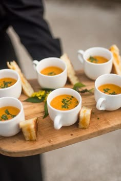 a wooden tray topped with cups of soup next to toasted bread on top of a table