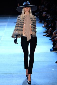 a woman walking down a runway wearing a black hat and jacket with fringes on it