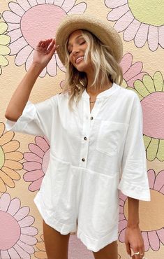 a woman standing in front of a flower wall wearing a hat and white shirt dress