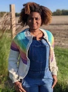 a woman standing in front of a fence wearing overalls and a colorful sweater with stripes on it