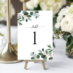 a table number card with greenery on it and white flowers in vases behind it