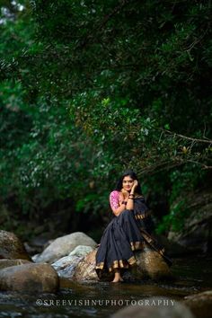 a woman sitting on top of a rock next to a river while talking on a cell phone