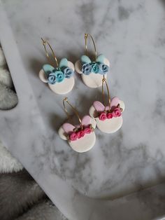 three earrings with pink and blue flowers are on a marble table next to a teddy bear