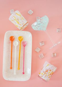 three plastic spoons and two glass cups on a pink surface with ice cubes