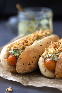 two hot dogs with toppings sitting on top of wax paper next to a jar