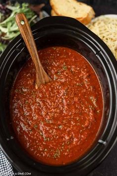 a wooden spoon in a slow cooker filled with marinara sauce next to bread