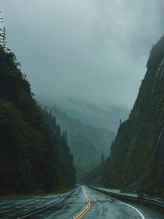 an empty road with mountains in the background and foggy skies above it on a rainy day