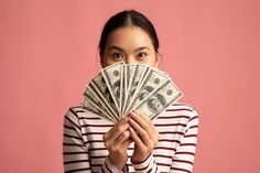 a woman covering her face with one hundred dollar bills in front of her face, against a pink background