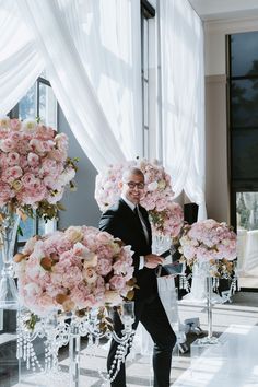 a man in a tuxedo standing next to tall pink flowers on a table