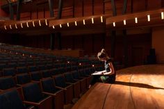 a woman sitting on the floor in front of an empty auditorium with blue chairs and wooden floors