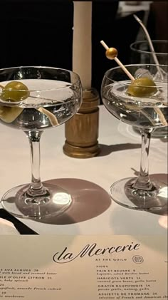 two martini glasses with olives in them sitting on a white tablecloth covered table