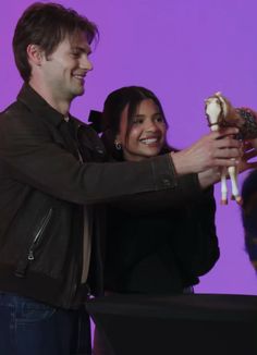 a man and woman standing next to each other in front of a purple background holding stuffed animals