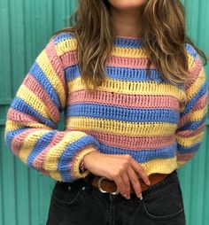 a woman with long hair wearing a multicolored sweater and black jeans standing in front of a green wall