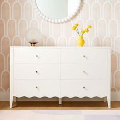 a white dresser sitting in front of a mirror on top of a wooden floor next to a vase with yellow flowers