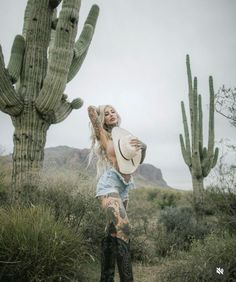 a woman standing in front of a cactus with her arms around her body and holding a hat